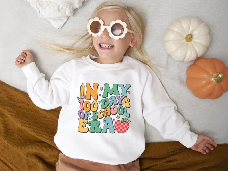 100th Day Shirt: Era A little girl wearing flower sunglasses is wearing a cream colored sweatshirt that says in my 100 days of school era.