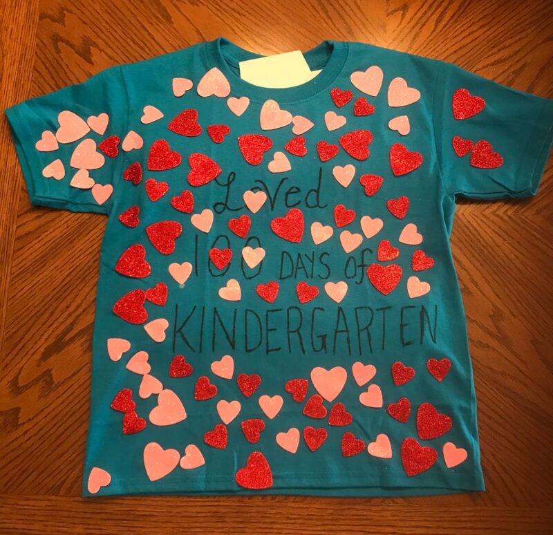100th Day Shirt: Hearts A teal t-shirt has red and pink heart stickers all over it. It says Loved 100 Days of Kindergarten in marker. (100th day of school shirt ideas)