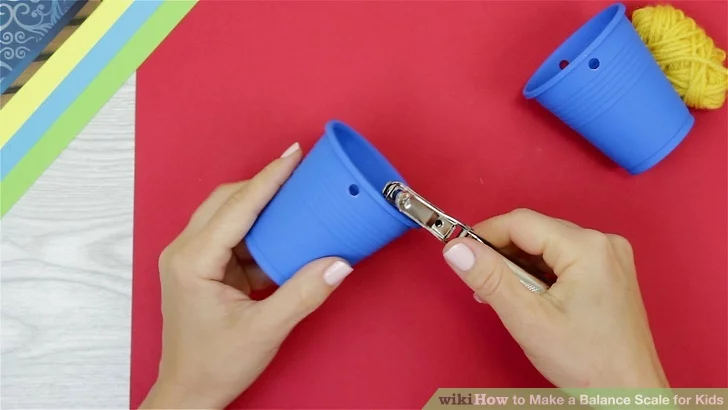 DIY Balance Scale Activity Hands are shown punching holes into small plastic cups (first grade science experiments)