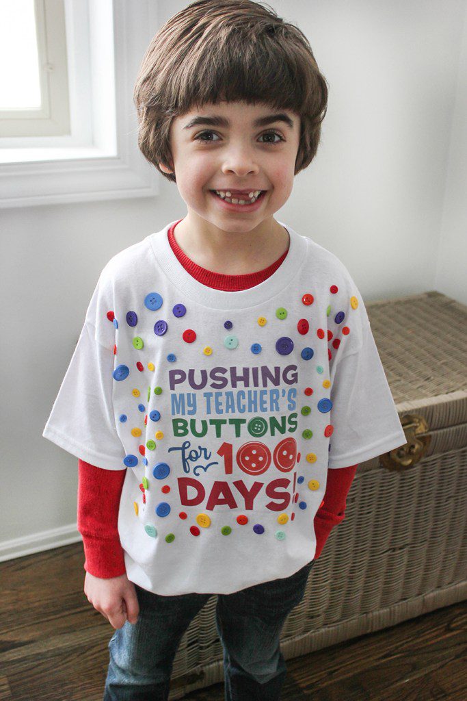 100th Day Shirt: Buttons 100th day of school shirt ideas include this one. A little boy is wearing a white t-shirt that says Pushing His Teacher's Buttons for 100 Days.
