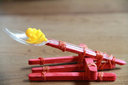 Catapult STEM Challenge popsicle sticks and elastics are stacked to create a catapult. A plastic spoon makes for the actual catapult piece (preschool science)