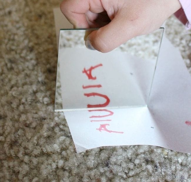 First Grade Science ExperimentsYou Young student's hand holding a mirror on a piece of paper with the letters A I U, showing symmetry (First Grade Science Experiments)