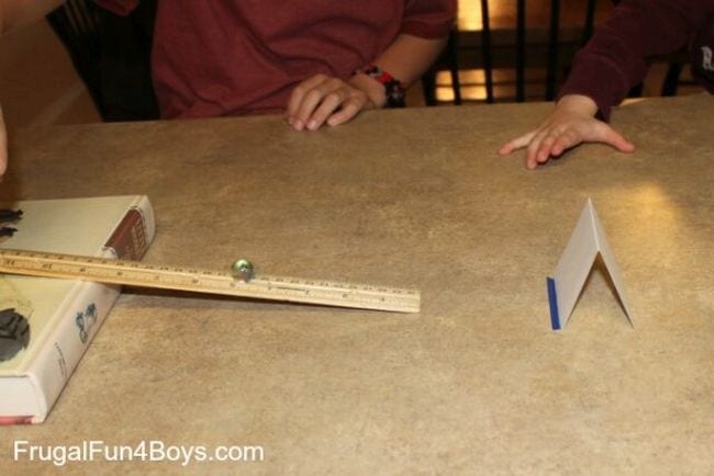 First Grade Science Experiments A ruler propped on one side on a flat book, with a marble rolling down it toward a folded index card (First Grade Science Experiments)