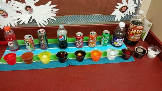 First Grade Science Experiments A series of plastic cups filled with varieties of soda, juice, and other liquids, with an egg in each