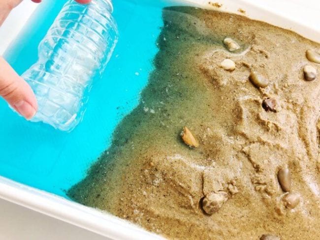Fourth Grade Science Coastal Erosion Plastic bin filled with sand, shells, and water to simulate a beach, with a hand holding a plastic bottle in the water (Fourth Grade Science)