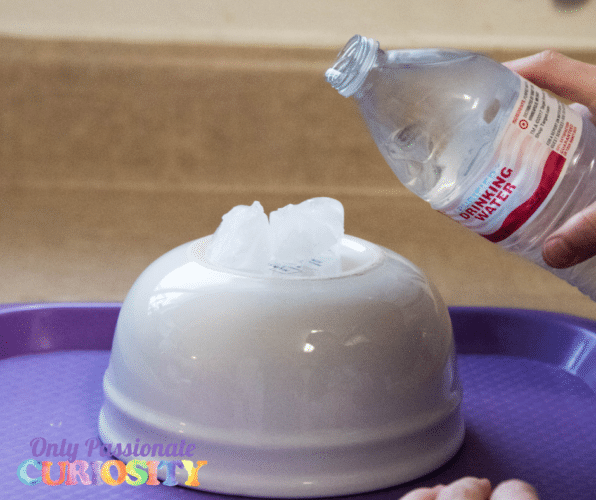 Instant Ice Experiment White ceramic bowl turned upside-down with ice cubes on top. A person is pouring water onto the ice from a plastic bottle.