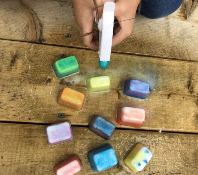 Kindergarten Science Activities Fizzing Ice Cubes Child's hands holding a spray bottle over colorful ice cubes on a wood surface