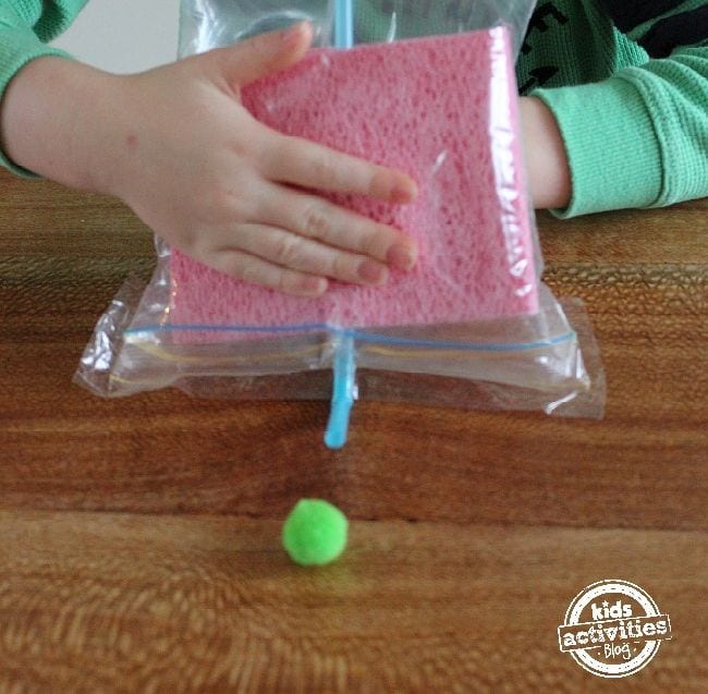 Preschool Science: Air Pressure Young student using a straw in a plastic bag with sponges to move a green pompom across a wood table