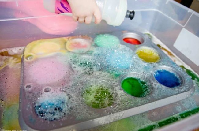 Preschool Science: Rainbow Baking Soda and Vinegar Preschool student squeezing a bottle of vinegar into muffin tin cups filled with baking soda and food coloring