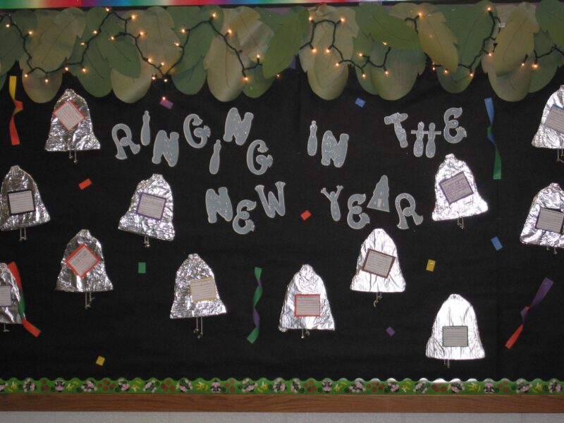 New Year Bulletin Ringing In A black bulletin board has silver bells with writing prompts on it. They are made from tin foil. Actual Christmas lights adorn the top and text reads ringing in the new year.