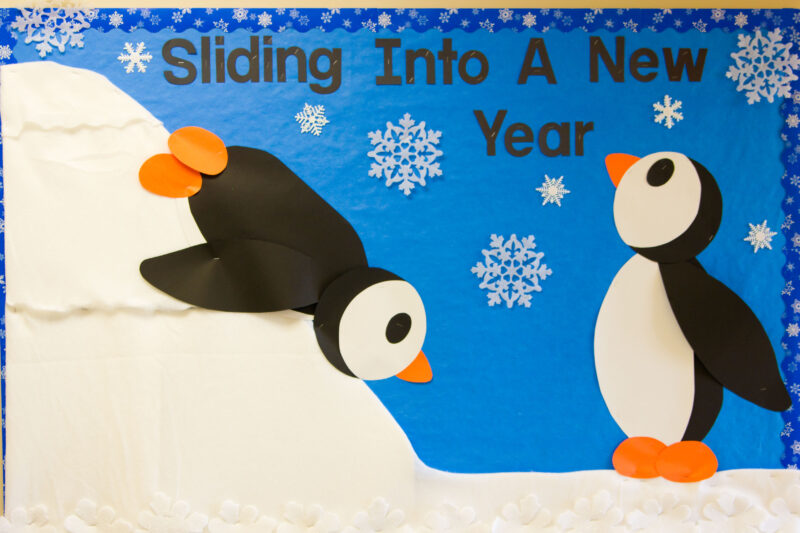 New Year Bulletin Penguins Sliding A bulletin board shows two penguins, one is sliding down a snowy hill on its stomach. Text reads,