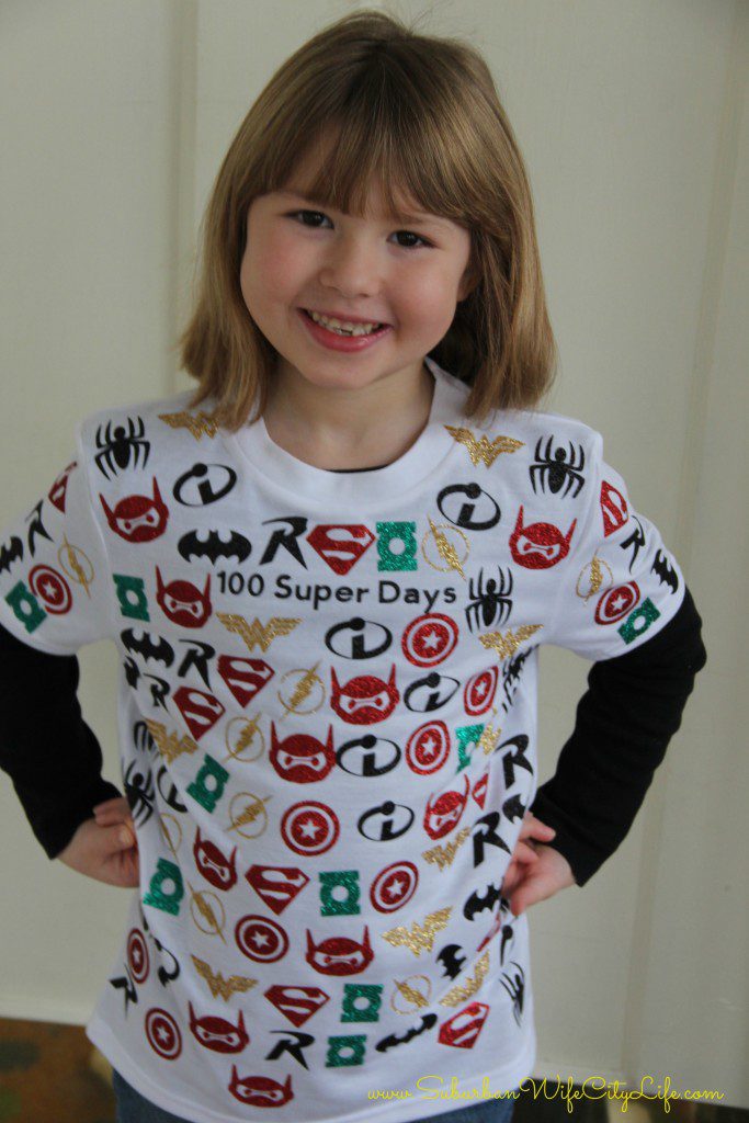 100th Day Shirt: Superhero A little girl with her hands on her hips is wearing a white t shirt that says 100 super days and there are many different superhero masks on it.