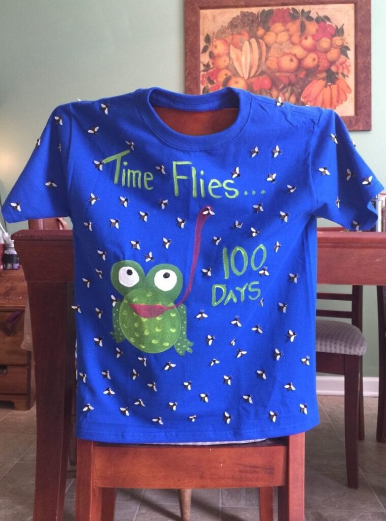 100th Day Shirt – Time Flies A blue homemade shirt says Time flies 100 days. It has a frog with a big tongue catching flies that are all over the shirt.