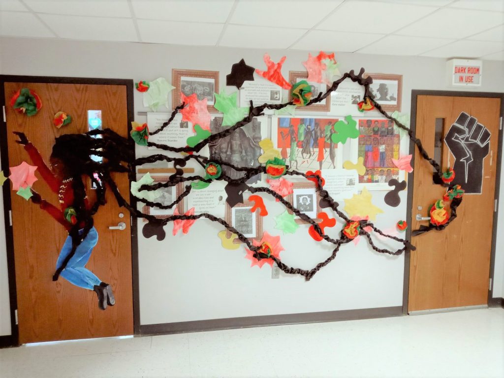 Black History Bulletin Board – Art A hallway is connected by two doorways. The door on the left has a side profile of a girl whose dreadlocks are running across the hallway into the second door. There is information on artists in the hallway.