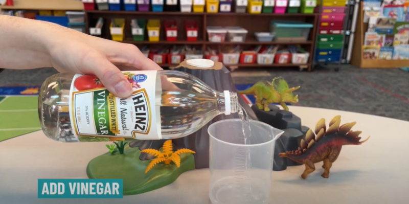 Volcano Step 3 A hand is shown pouring vinegar into a plastic measuring cup in this step of a baking soda volcano.
