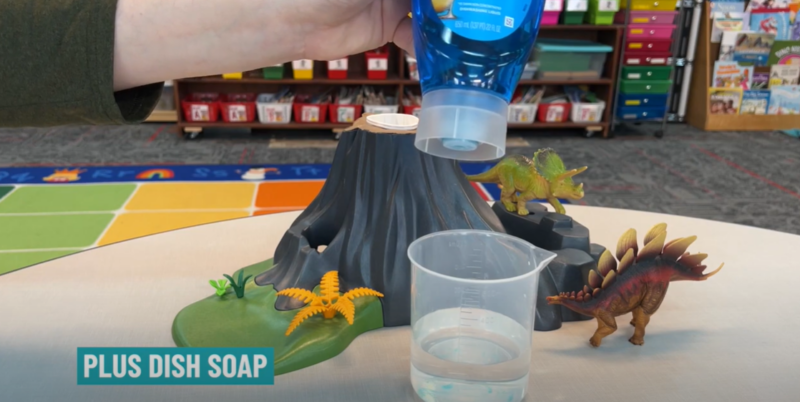 Volcano Step 4 Text reads Plus Dish Soap. This step of a baking soda volcano shows dish soap being poured into a plastic measuring cup.