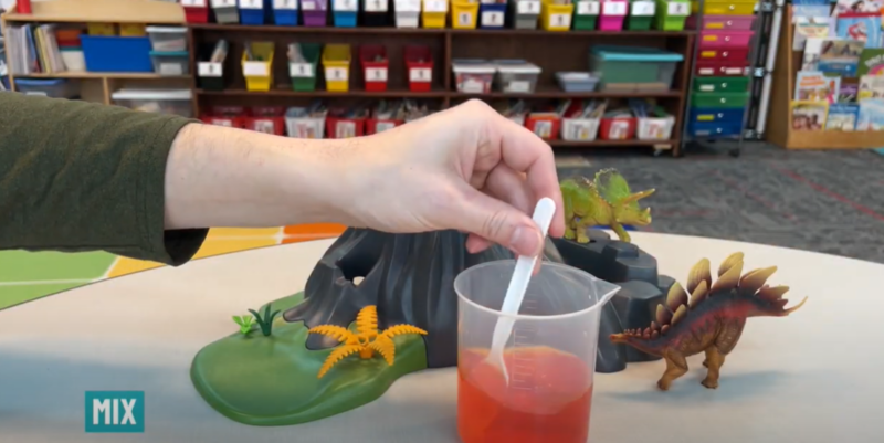 Volcano Step 6 Text reads mix and a hand is seen mixing red liquid in a plastic measuring cup with a spoon.