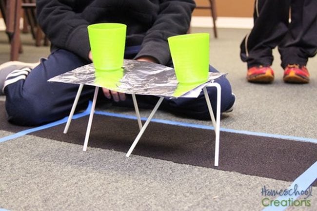 8th Grade Science Homeschool Creations Student showing a foil platform balanced on drinking straws, holding two yellow plastic cups of water