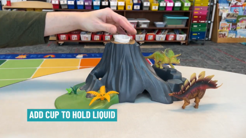 Volcano Step 1 A hand is seen placing a paper cup in the top of a volcano mold. Text reads Add cup to hold liquid.