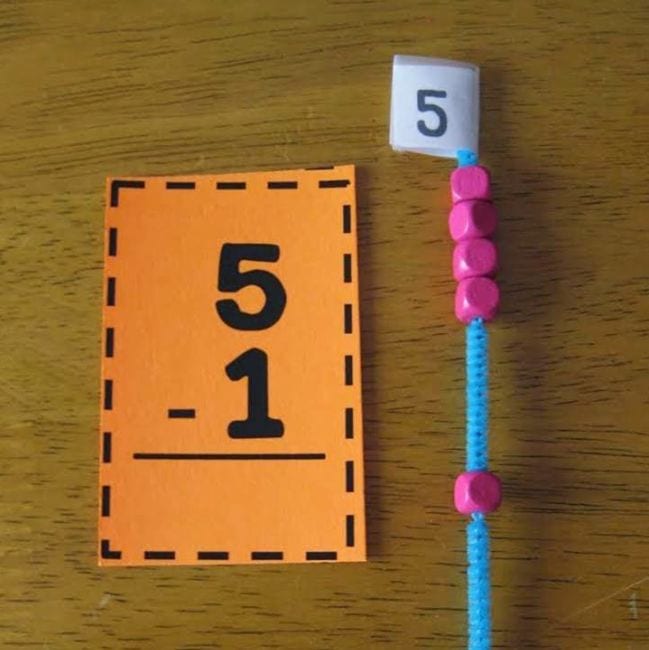 bead-subtraction A subtraction problem on a bright orange card next to 5 pink beads on a blue pipe cleaner