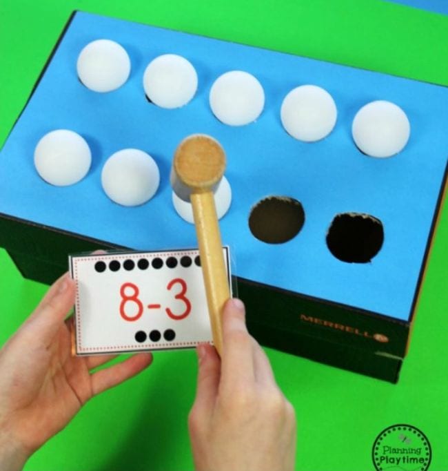 whack-a-ball-math-activity A colored paper-covered shoe box with holes cut on the top. In each of the holes is a ping pong ball. A hand holds a mallet and a math problem