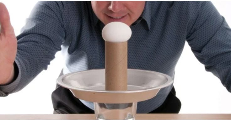 Eighth Grade Science Intertia Egg Drop Egg on top of a toilet paper tube, standing on a plate on a glass of water, with a man ready to hit the plate