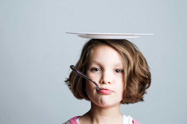 Plate on Head A young girl is shown from the neck up with a plate on her head and a spoon in her mouth.