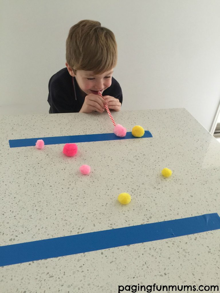 Pom Pom Blowing Staw Minute to win it games include this one where a little boy blows yellow and pink pom poms across a table by blowing into a straw.