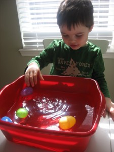 sinkorfloat A little boy stands in front of a red tub filled with water. There are various plastic Easter eggs inside.