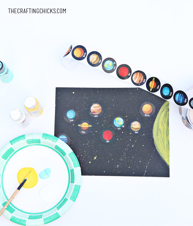 solarsystemstickers A plate has yellow paint on it with a paintbrush. A solar system scene has been painted and planet stickers have been stuck to it (solar system projects)