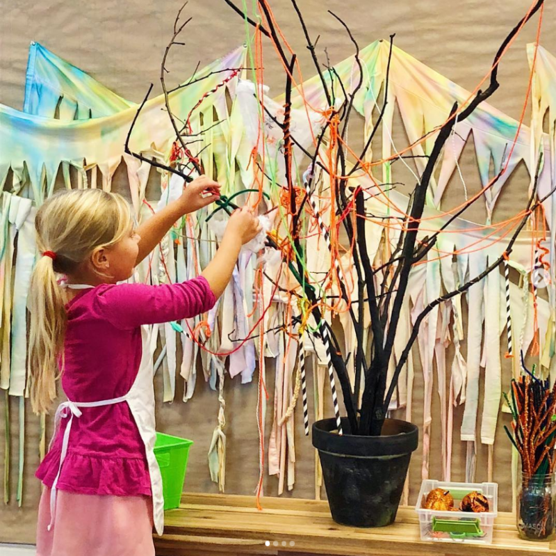 A little girl stands before a fake tree with a lot of branches that have been decorated with ribbons, beads, etc.