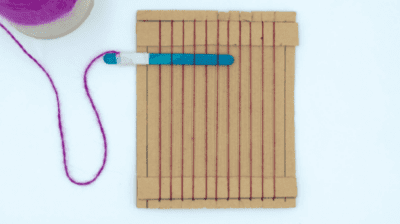 learn-to-weave A piece of cardboard with strings strung in rows vertically and a popsicle stick with yarn tied to the end woven into the top section of the strings as an example of summer crafts for kids