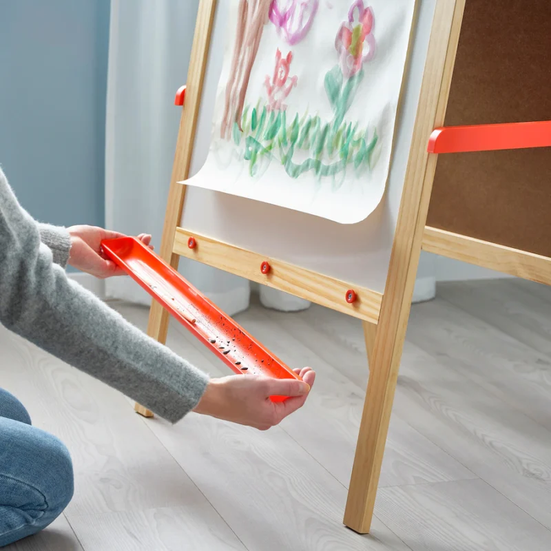 An easel is shown with a painting of lowers on it. Hands are shown removing a basin from the bottom of the easel in this example of Ikea classroom.