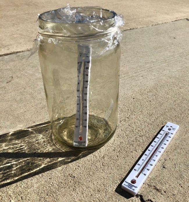 Outdoor Science Greenhouse Effect Glass jar covered in plastic wrap with a thermometer inside, next to a thermometer lying on the ground (Outdoor Science)