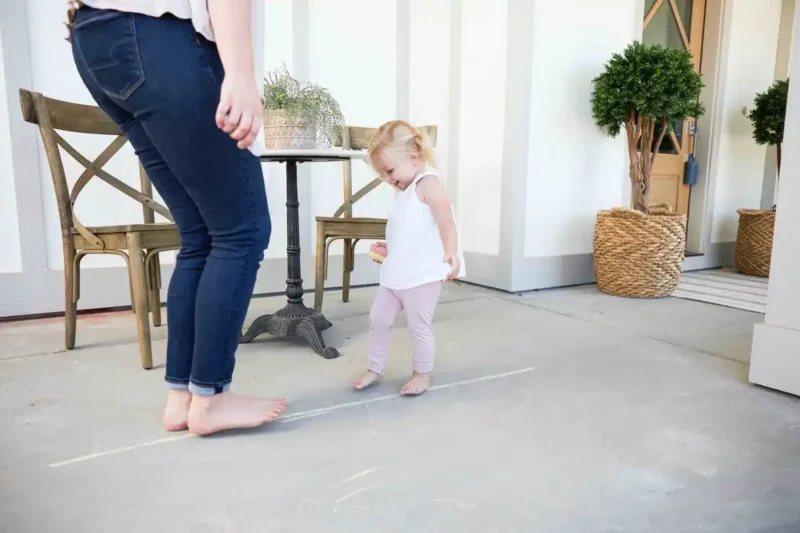Toddler_Obstacle_Course-1 A woman from the waist up is seen facing a small toddler girl in a living room.