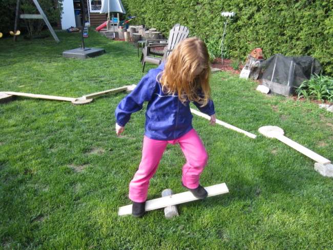 DIY Obstacle Courses for Kids: Loose Parts Child balancing on a plank of wood over a round piece, as part of diy obstacle courses for kids