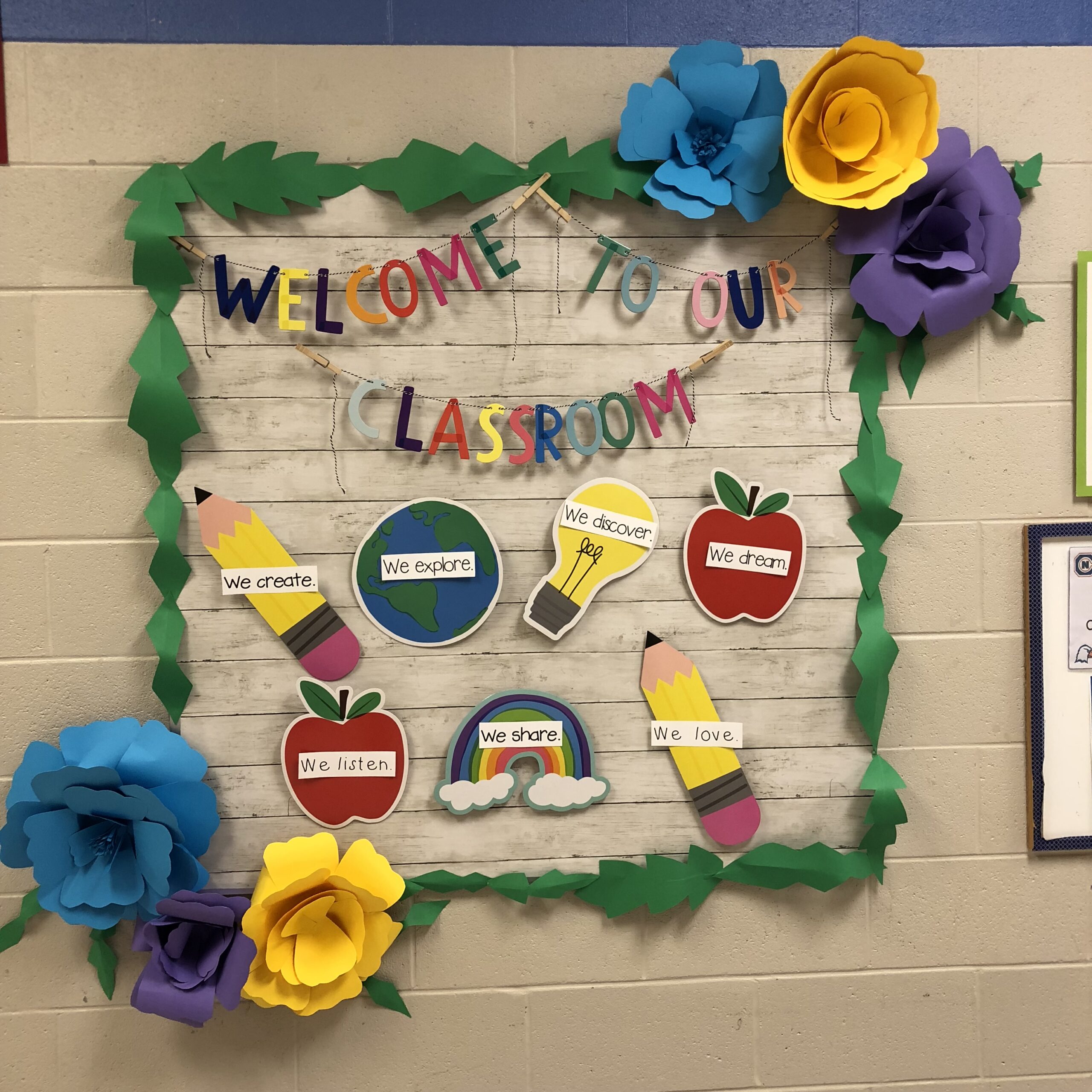 Flowers-Hilary-Statum A sign says Welcome to Our Classroom. The corners of the bulletin board have large oversize 3D flowers.