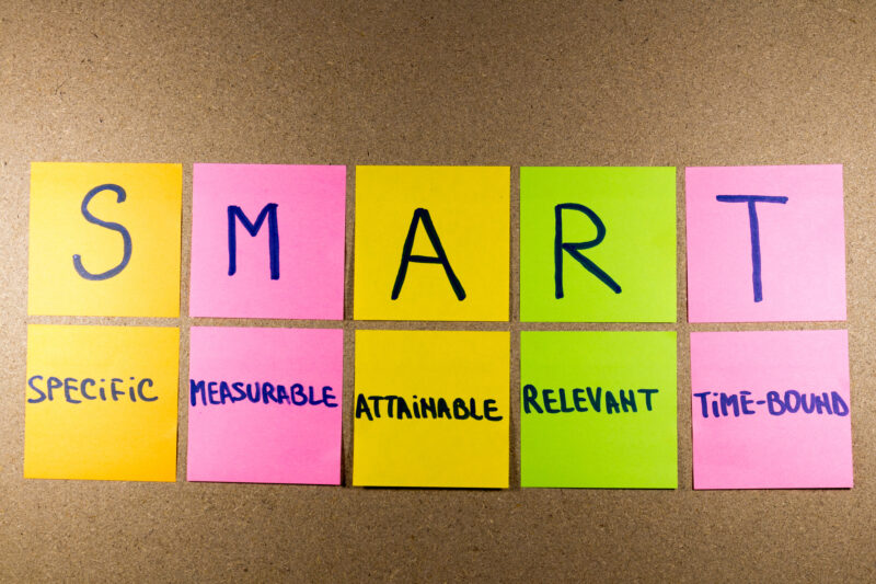 SMART Goals Set goals SMART (specific, measurable, attainable, recorded, timely) colorful sticky notes on cork bulletin board.