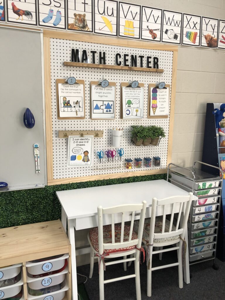 Math-Center-Hilary-Statum A corkboard says Math Center at the top and has several clipboards with different math assignments on it. A desk with two chairs is underneath it.