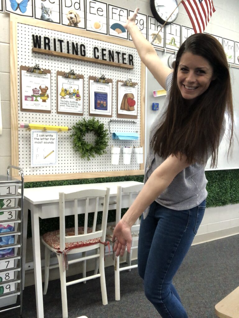 Writing-Center-Hilary-Statum A teacher stands with her arms out showing off a board that says Writing Center. There are several clipboards attached to it.