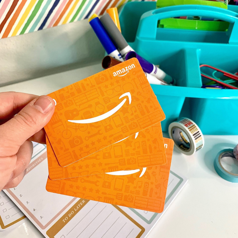 Amazon Gift Cards Hand holding two Amazon gift cards above a desk with art supplies.
