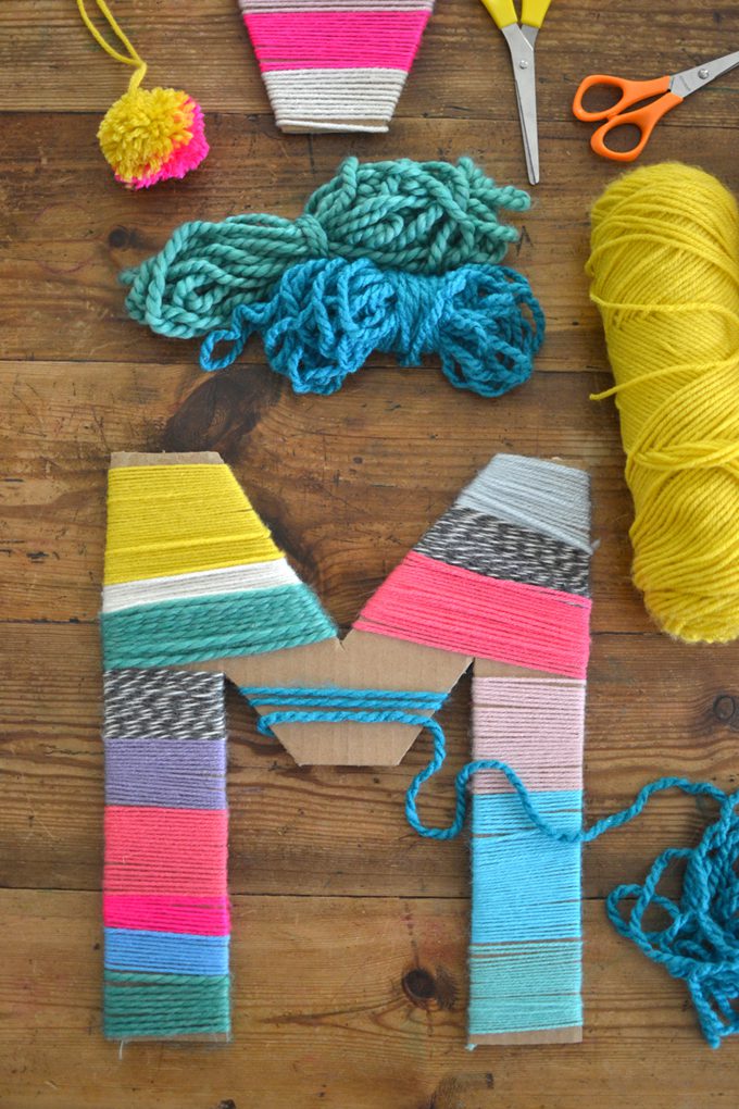Yarn Wrapped Letter A letter M made of cardboard is wrapped with different colored yarn. Several rows of yarn are in the background.