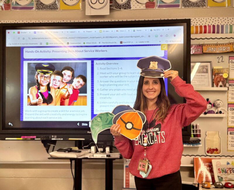image of white female teacher holding different community hats Image of white female teacher standing in front of lesson on a monitor and holding up a picture of a police person's hat